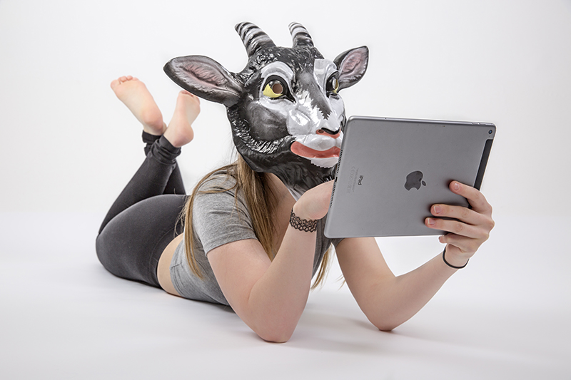 Das Bild zeigt eine Frau mit Ziegenkopf die auf einem Tablet scrollt und auf dem Boden liegt Das Bild zeigt eine Frau mit Ziegenkopf die auf einem Tablet scrollt und auf dem Boden liegt